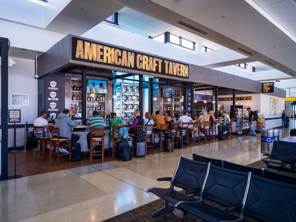The American Craft Tavern at John Glenn Columbus International Airport, featuring a large illuminated sign above an open bar and dining area. Travelers sit on barstools and at high-top tables, with shelves of beer taps and bottles visible behind the bar. Bright natural light streams in from tall windows above the space, and airport seating is visible in the foreground.