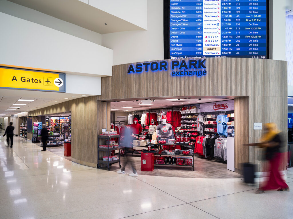 The Astor Park Exchange store at John Glenn Columbus International Airport, featuring Ohio State–themed apparel, gifts, and souvenirs displayed at the entrance. The storefront has a textured wood-paneled design with a bright blue sign. Travelers walk past in the concourse, and a large flight information display hangs above showing departure times.