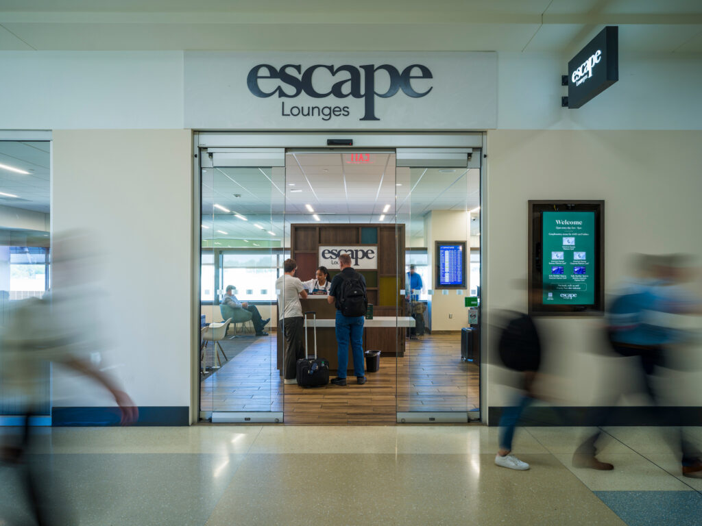 Entrance to the Escape Lounge at John Glenn Columbus International Airport, shown through glass sliding doors with the lounge’s logo above. A staff member assists two travelers at the check-in desk, while other passengers sit inside near large windows. A digital welcome sign is mounted on the wall to the right of the entrance. Several blurred travelers walk past in the foreground.