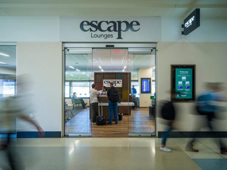 Entrance to the Escape Lounge at John Glenn Columbus International Airport, shown through glass sliding doors with the lounge’s logo above. A staff member assists two travelers at the check-in desk, while other passengers sit inside near large windows. A digital welcome sign is mounted on the wall to the right of the entrance. Several blurred travelers walk past in the foreground.