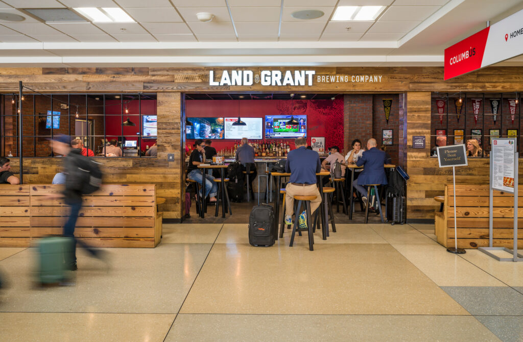The Land-Grant Brewing Company restaurant at John Glenn Columbus International Airport, featuring a rustic wood exterior and an open entrance leading to a bar area. Travelers sit at high-top tables and the bar, with multiple TVs displaying sports above the drink selection. A “Please Seat Yourself” sign stands near the entrance, and a few passengers walk by in a motion blur in front of the restaurant.