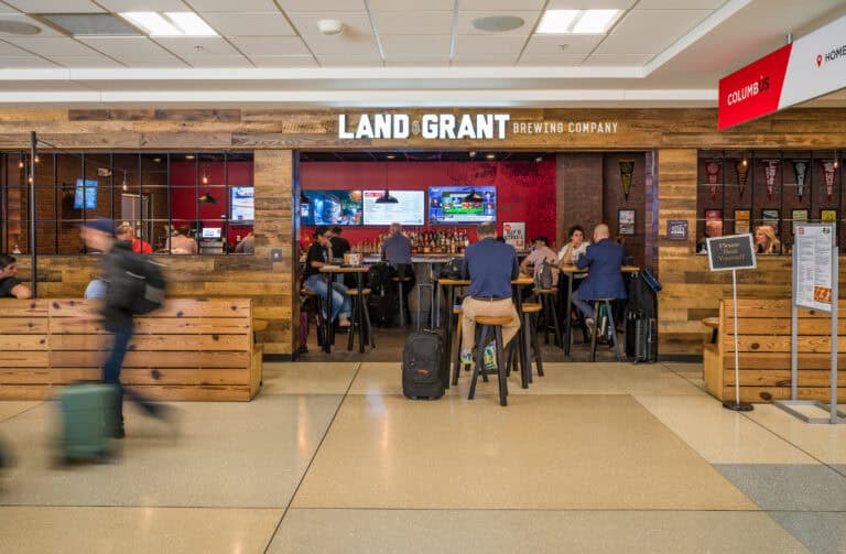 The Land-Grant Brewing Company restaurant at John Glenn Columbus International Airport, featuring a rustic wood exterior and an open entrance leading to a bar area. Travelers sit at high-top tables and the bar, with multiple TVs displaying sports above the drink selection. A “Please Seat Yourself” sign stands near the entrance, and a few passengers walk by in a motion blur in front of the restaurant.
