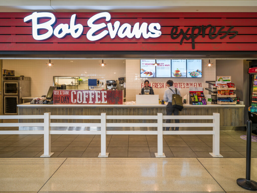 The Bob Evans Express counter at John Glenn Columbus International Airport, featuring a bright red and white illuminated sign above the ordering area. A customer stands at the register while a staff member takes their order. Digital menu boards display breakfast items, salads, and wraps. A display of snacks and bottled drinks is visible to the right, and a white railing separates the counter from the walkway.