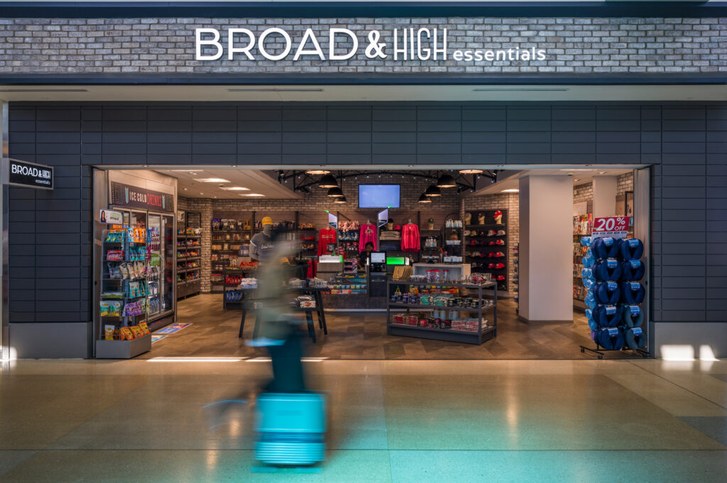 The Broad & High Essentials store at John Glenn Columbus International Airport, with a brick-accented storefront and bright illuminated sign. Inside, shelves display snacks, drinks, travel essentials, Ohio-themed apparel, and souvenirs. A traveler with a rolling suitcase walks past the entrance in a motion blur.