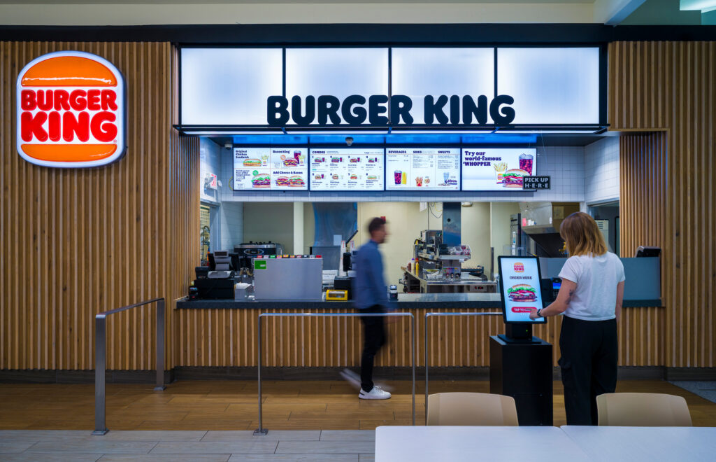 The Burger King counter at John Glenn Columbus International Airport, featuring a bright illuminated sign and digital menu boards displaying burgers, combos, and drinks. A traveler uses a self-order kiosk in the foreground, while another person walks past the counter. The space has modern wood paneling and a clean, open design.