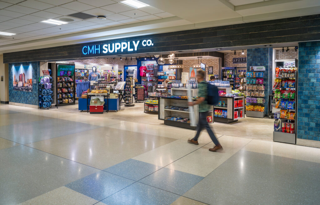 The CMH Supply Co. store at John Glenn Columbus International Airport, with a bright illuminated sign above the entrance. The shop displays a wide range of snacks, drinks, travel essentials, apparel, and Columbus-themed souvenirs. Shelves and tables are filled with colorful merchandise, and a blurry traveler walks past the storefront in the foreground.
