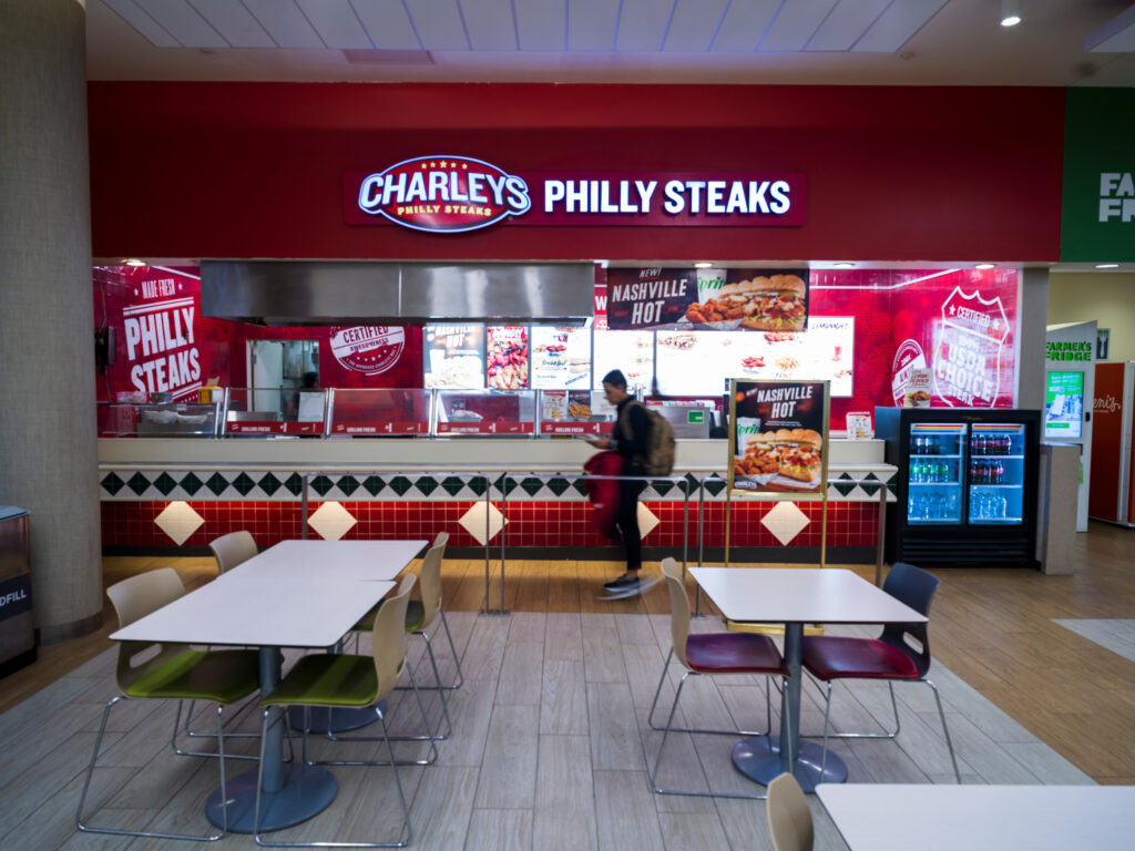 The Charleys Philly Steaks counter at John Glenn Columbus International Airport, featuring a bright red sign and menu boards showcasing sandwiches and combo meals. A customer walks past in front of the counter, and nearby tables and chairs sit empty in the dining area. A refrigerated case with bottled drinks is located to the right of the counter.