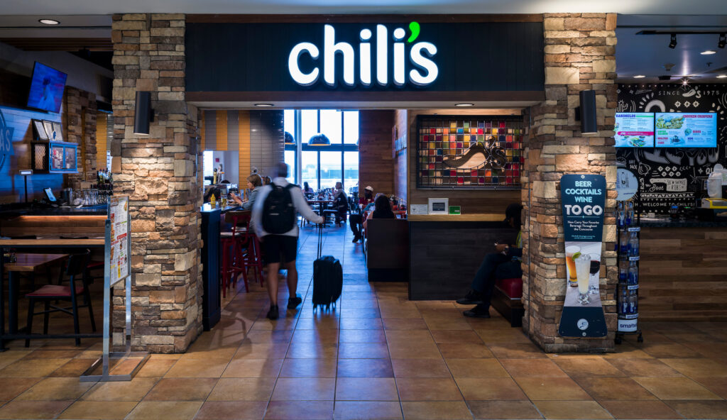 The entrance to Chili’s at John Glenn Columbus International Airport, framed by stone pillars and a bright illuminated Chili’s sign. Inside, diners sit at tables and the bar, and a traveler with a rolling suitcase walks into the restaurant. A colorful chili pepper artwork decorates the wall on the right, along with a sign promoting to-go drinks.