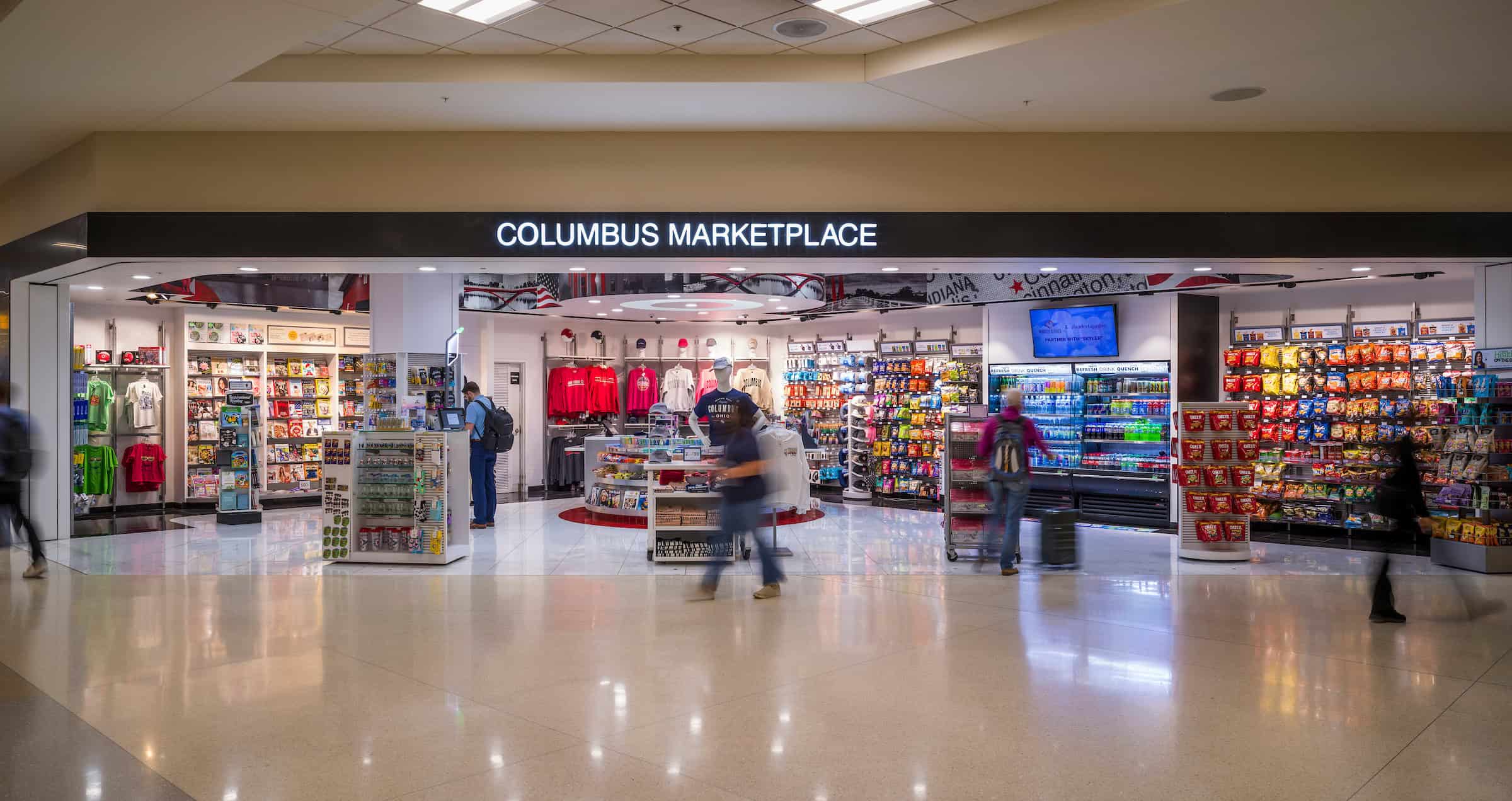The Columbus Marketplace store at John Glenn Columbus International Airport, brightly lit with shelves of snacks, drinks, magazines, souvenirs, and Ohio-themed apparel. Several travelers browse inside while others walk past in a motion blur. The store’s name appears in large illuminated letters above the entrance.