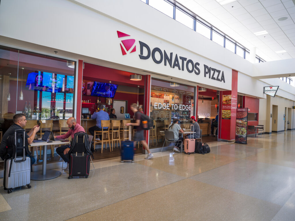 The Donatos Pizza restaurant at John Glenn Columbus International Airport, with a bright white exterior sign and large windows looking into the dining area. Travelers sit at tables and the bar inside, while others with luggage walk by in the concourse. Multiple TVs above the bar display news and sports, and signage promotes Donatos’ signature “edge to edge” toppings.