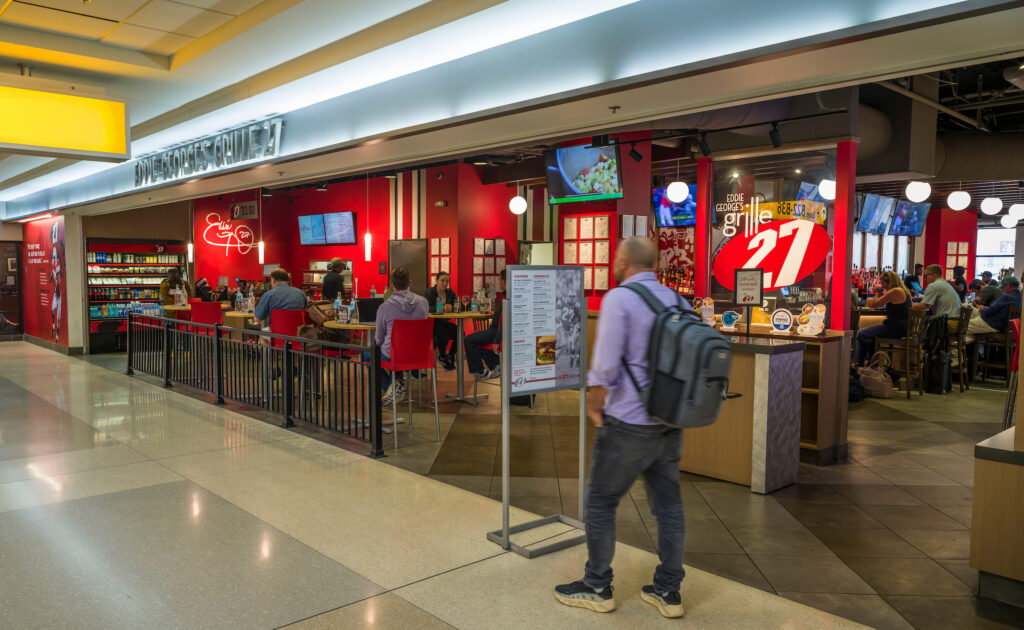 Eddie George’s Grille 27 at John Glenn Columbus International Airport, with a bright red interior, bar seating, and multiple TVs showing sports. Guests dine at tables near the entrance, and a large “27” logo is displayed on a glass panel. A traveler with a backpack walks past the restaurant, which also features a grab-and-go cooler stocked with drinks and snacks.