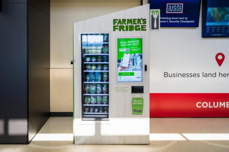A Farmer’s Fridge vending machine at John Glenn Columbus International Airport, styled in light wood with green accents. The machine displays rows of fresh grab-and-go items like salads, bowls, and snacks in clear jars. A touchscreen on the right promotes the Farmer’s Fridge app and payment options. A small slot below the screen is labeled “Recycle jars here.” Bright sunlight casts shadows across the floor.