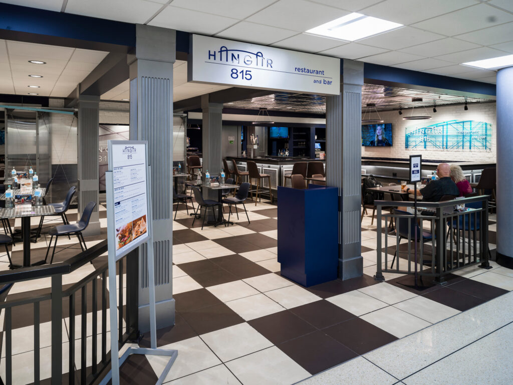 The Hangar 815 restaurant and bar at John Glenn Columbus International Airport, featuring a modern aviation-inspired design with blue and gray accents. Tables and seating fill the dining area, with TVs mounted above the bar. A neon-style wall sign displays “Hangar 815,” and a menu stand sits at the restaurant entrance. Travelers dine at several tables inside.