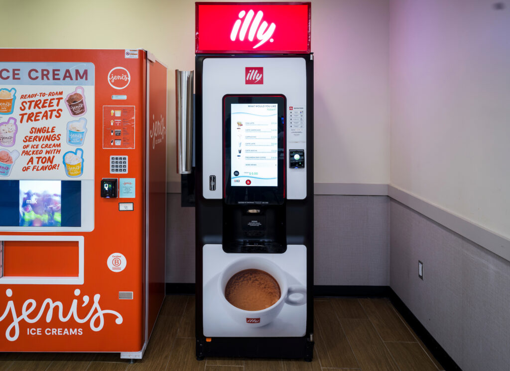 An Illy coffee vending machine at John Glenn Columbus International Airport, featuring a tall digital touchscreen displaying drink options such as lattes and cappuccinos. The machine has a red illuminated Illy sign at the top and a large image of a cup of coffee at the bottom. A Jeni’s ice cream vending machine is partially visible to the left.