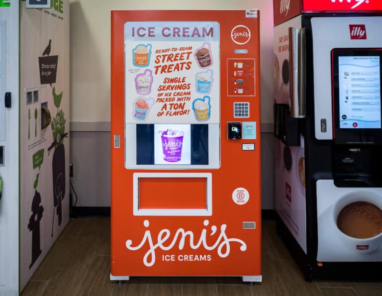 A bright orange Jeni’s Ice Cream vending machine at John Glenn Columbus International Airport, decorated with illustrated ice cream scoops and text promoting single-serving “street treats.” A digital window displays available flavors, and a card reader and keypad are located on the right side of the machine.