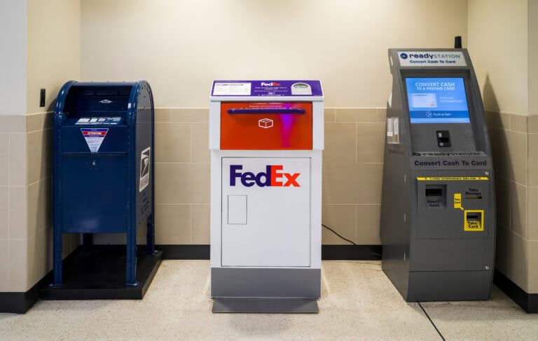 A USPS blue mail drop box, a white and orange FedEx drop box, and a gray ReadyStation machine for converting cash to a prepaid card are lined up against a beige tiled wall at John Glenn Columbus International Airport.