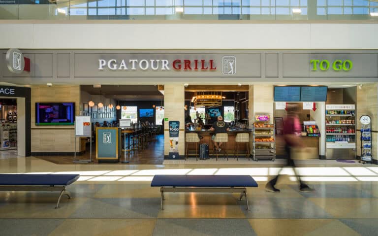 The PGA Tour Grill at John Glenn Columbus International Airport, featuring a wide entrance with illuminated signage for both the main restaurant and a “To Go” section. Travelers sit at the bar inside, and shelves of snacks, drinks, and grab-and-go items line the right side. Warm lighting and golf-themed decor create a welcoming atmosphere, while a traveler walks past in the foreground in motion blur.