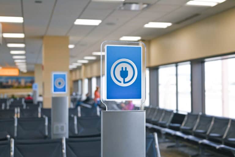 Power towers stand above the seats in a gate area at the airport. The tops are lit up blue with a power cord icon on the front.