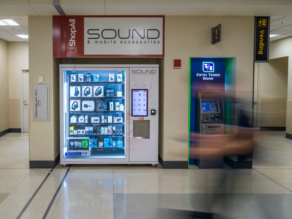 A SOUND & mobile accessories vending machine at John Glenn Columbus International Airport, stocked with headphones, chargers, and other electronics. The machine is brightly lit with a touchscreen on the right side. Next to it is a Fifth Third Bank ATM with blue and green accent lighting. A traveler walks by in motion blur.