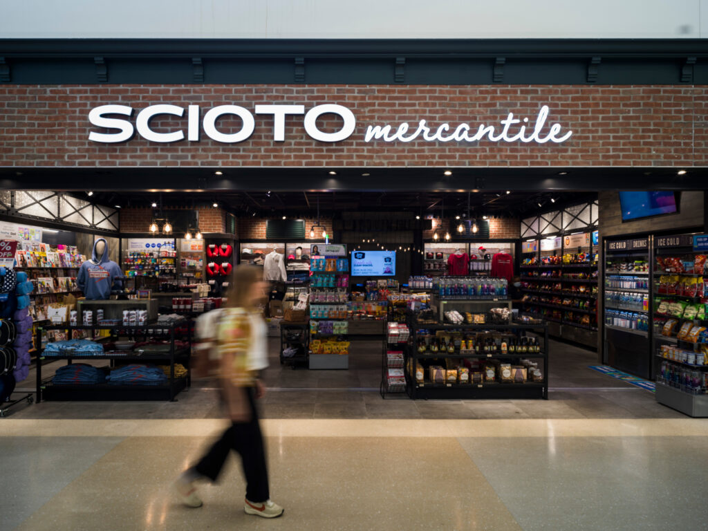 The Scioto Mercantile store at John Glenn Columbus International Airport, featuring a brick storefront with a bright illuminated sign. Inside, shelves display snacks, drinks, apparel, souvenirs, and travel essentials. A traveler walks past in the foreground in a slight motion blur.