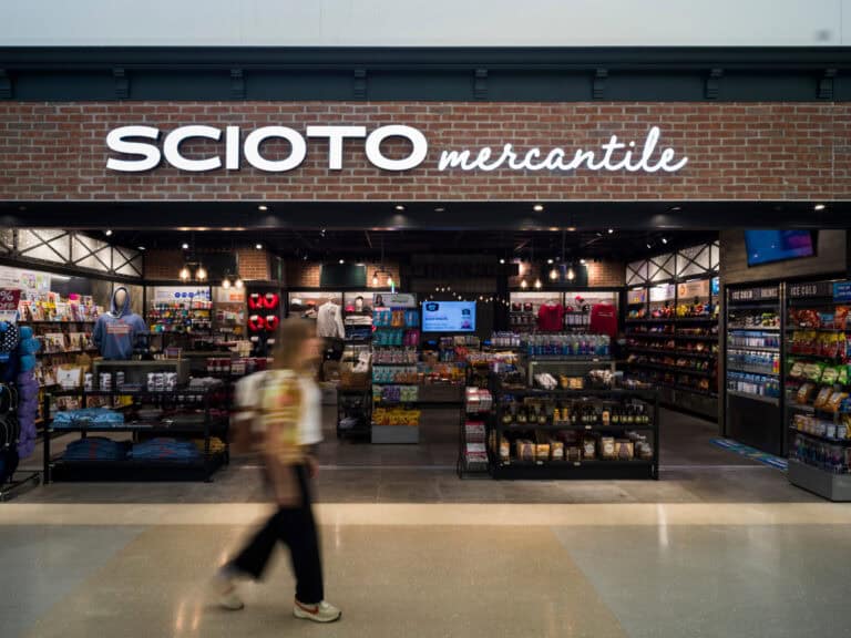 The Scioto Mercantile store at John Glenn Columbus International Airport, featuring a brick storefront with a bright illuminated sign. Inside, shelves display snacks, drinks, apparel, souvenirs, and travel essentials. A traveler walks past in the foreground in a slight motion blur.