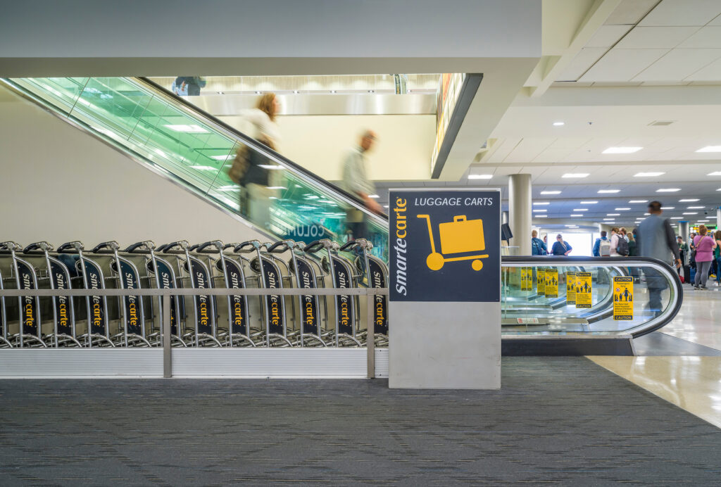 A row of SmarteCarte luggage carts lined up near an escalator inside John Glenn Columbus International Airport. A large sign with a yellow luggage cart icon reads “SmarteCarte Luggage Carts.” Travelers ride the escalator in motion blur above, and more passengers walk through the terminal in the background.