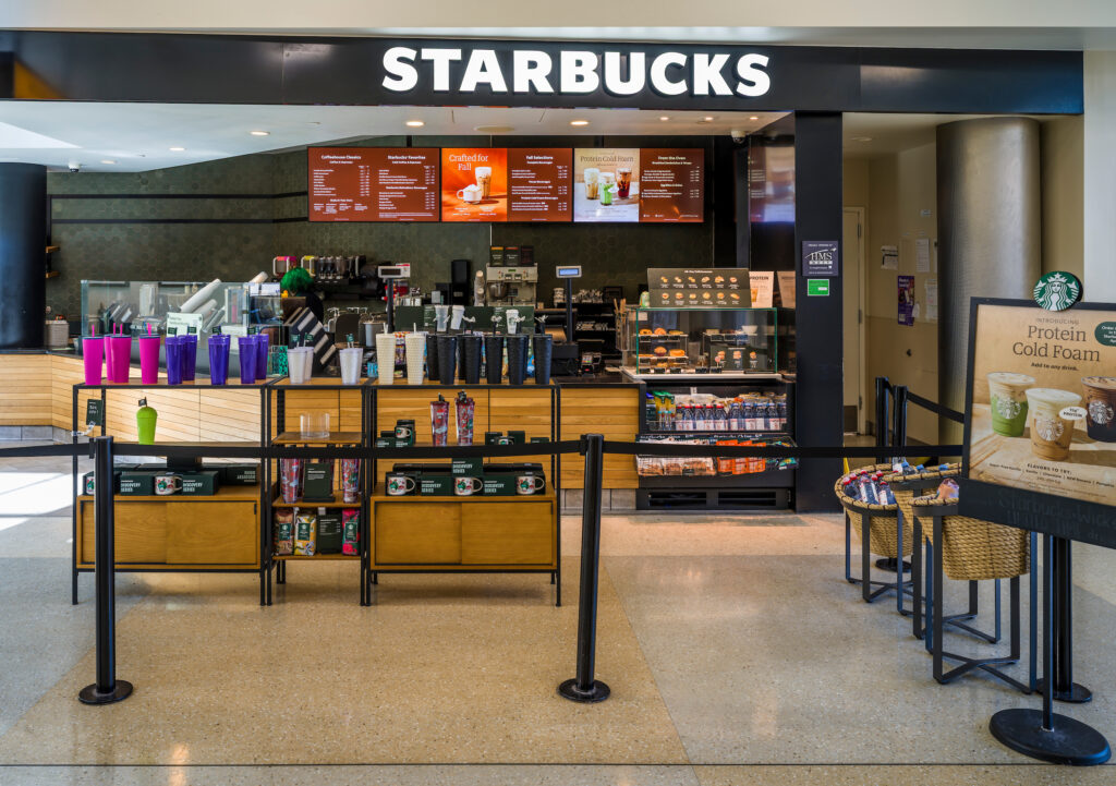 The Starbucks location at John Glenn Columbus International Airport, featuring a bright illuminated sign and a front display of colorful reusable cups and merchandise. Behind the counter, baristas prepare drinks, with menu boards showing seasonal and standard offerings. A refrigerated case displays bottled beverages and snacks near the register.