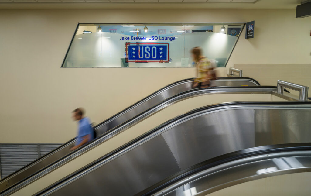 Two travelers ride escalators in opposite directions at John Glenn Columbus International Airport, seen in motion blur. Above them, a glass-walled USO space displays the sign “Jake Brewer USO Lounge” with the USO logo.