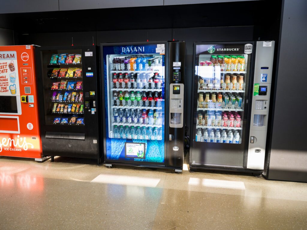 A row of vending machines at John Glenn Columbus International Airport, including a Jeni’s Ice Cream machine on the left, a black snack vending machine stocked with chips and candy, a blue Dasani machine filled with bottled water and soft drinks, and a Starbucks-branded machine offering bottled coffees and juices. Sunlight reflects across the floor in front of the machines.