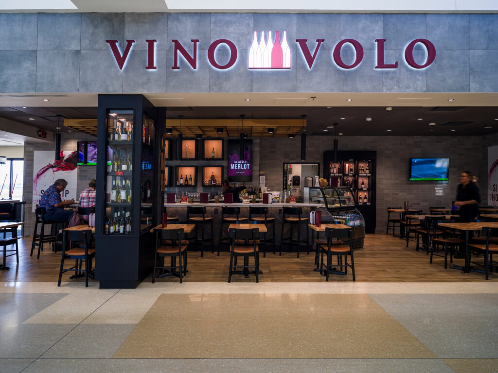 Vino Volo wine bar at John Glenn Columbus International Airport, featuring a sleek gray exterior with a glowing red sign. Inside, a central bar displays wine bottles and a glass case with food items. Wooden tables and chairs fill the dining area, where a few travelers are seated. A staff member walks past on the right, and a TV screen hangs on the back wall.