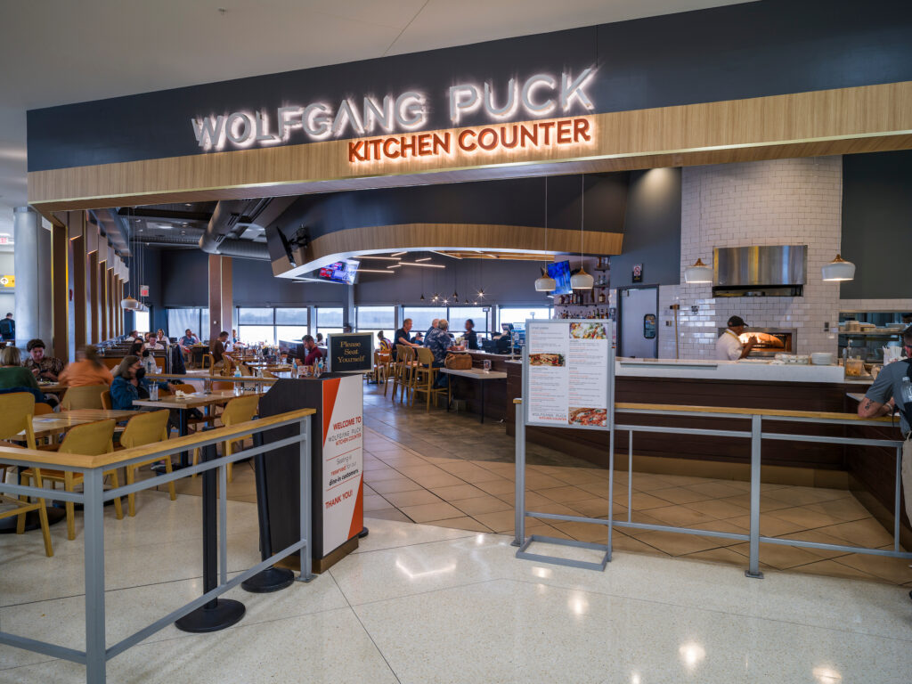 The Wolfgang Puck Kitchen Counter restaurant at John Glenn Columbus International Airport, with a glowing overhead sign and an open dining area filled with travelers seated at tables. A chef works at a wood-fired oven on the right side, and a “Please Seat Yourself” sign stands near the entrance. Large windows in the back provide natural light throughout the space.