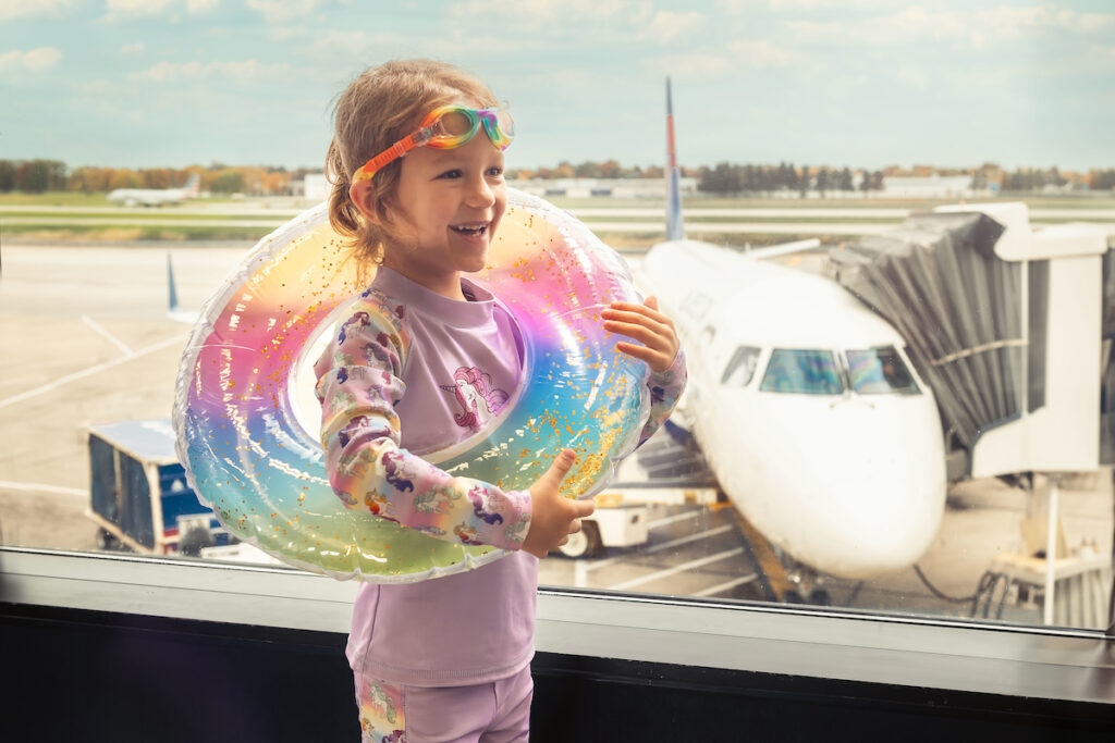 A little girl wearing swimming goggles on her head and a multi-color inner tube around her waist stands by an airport window, smiling off-camera. A white airplane sits at a gate behind her.