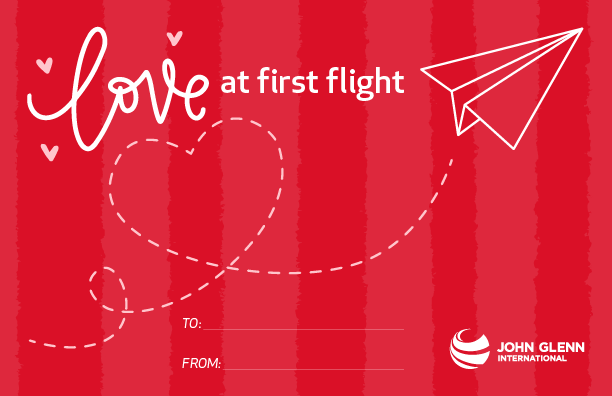 A Valentine's Day card with a paper airplane flying across the card, with a dotted line in the shape of a heart behind it. Text reads, "Love at first flight."