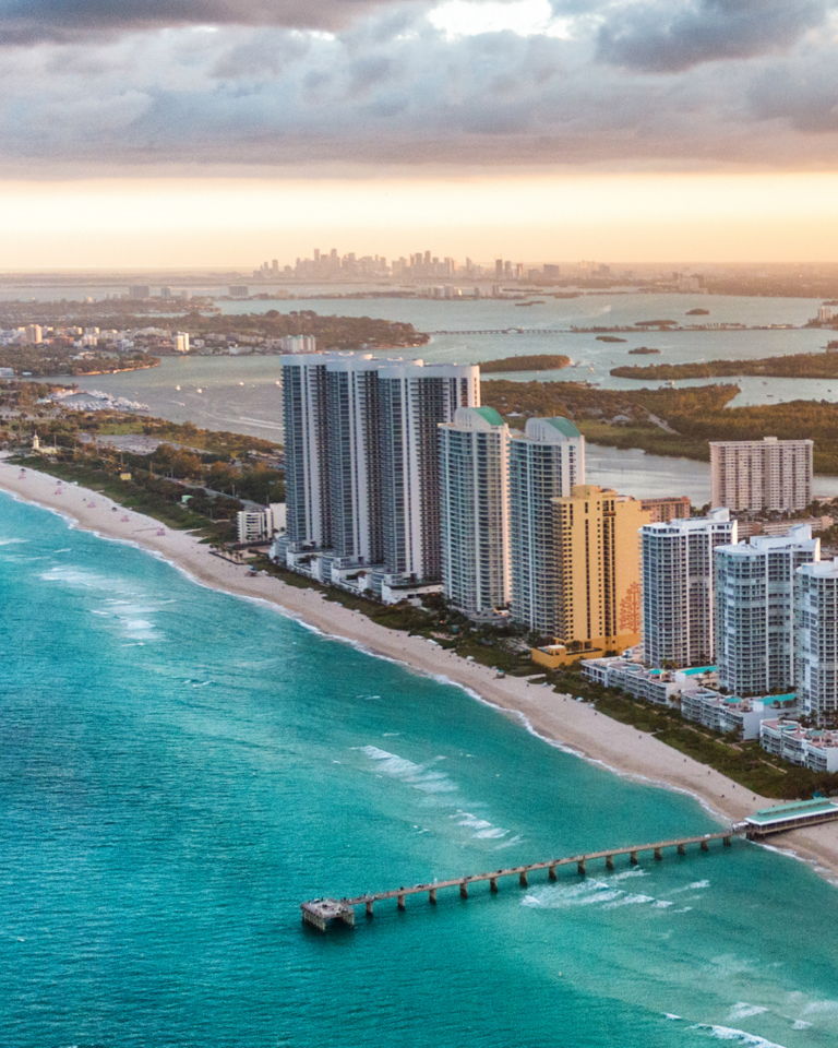 Miami Beach from above at sunset. Buildings stand along the shoreline, as well as in the distance.
