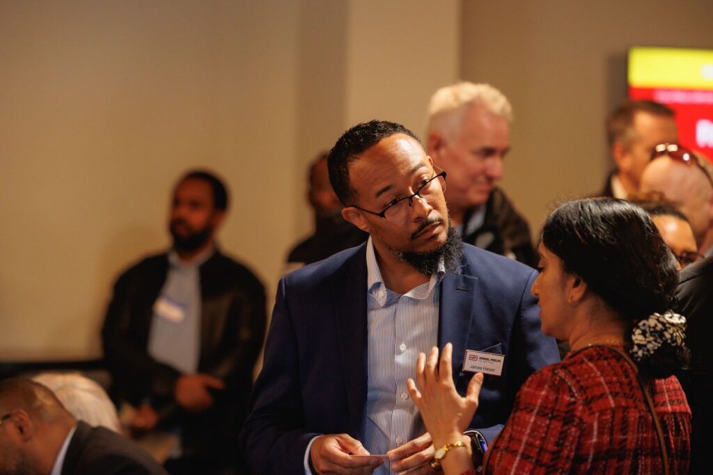 Two individuals speak to one another as more people mingle in the background. They're indoors at a networking event.