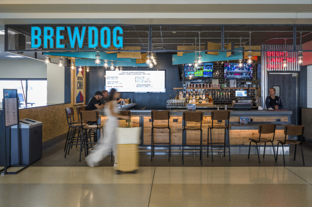 The BrewDog bar inside John Glenn Columbus International Airport, featuring a bright blue BrewDog sign above a wood-and-metal bar with high-top seating. A bartender stands behind the counter, and two guests sit at the bar. Multiple TVs display sports, and shelves of beer cans and bottles line the back wall. A traveler with rolling luggage walks past in a motion blur.