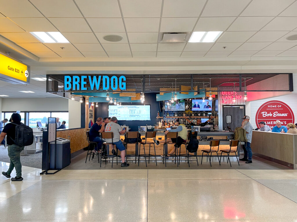 BrewDog storefront at John Glenn International Airport. To the left, an American Airlines gate crew stands at their computer. In the middle, passengers sit at the barstools at BrewDog enjoying a drink before their flight. To the right is the corner of Bob Evans Express' wall.