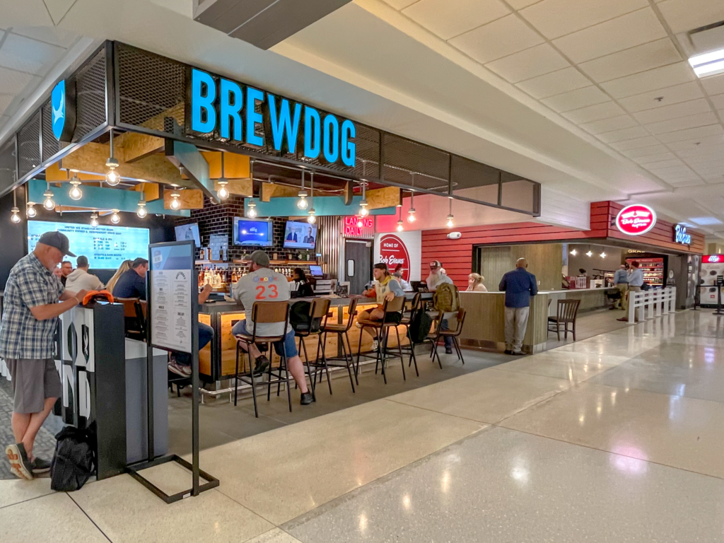 BrewDog's storefront at John Glenn International Airport. Several passengers sit at barstools at the bar, enjoying drinks before their flights. Bob Evans Express is seen in the background.