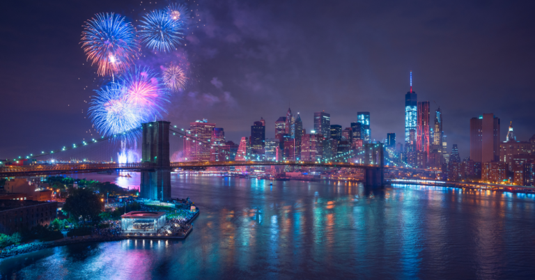 Bright red and blue fireworks light up the sky over the Brooklyn Bridge. The skyline of New York City is lit up in the background.
