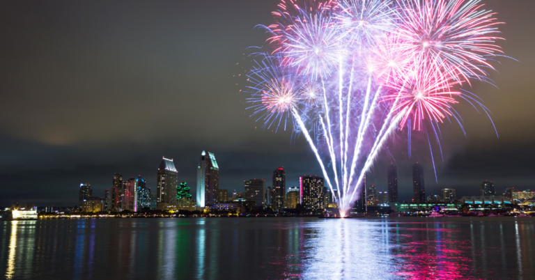 Bright red, white, and blue fireworks shoot from a barge on the water over San Diego at night.