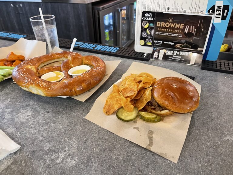 A large soft pretzel sits on a plate with two dipping sauces. Next to it, a burger with potato chips.