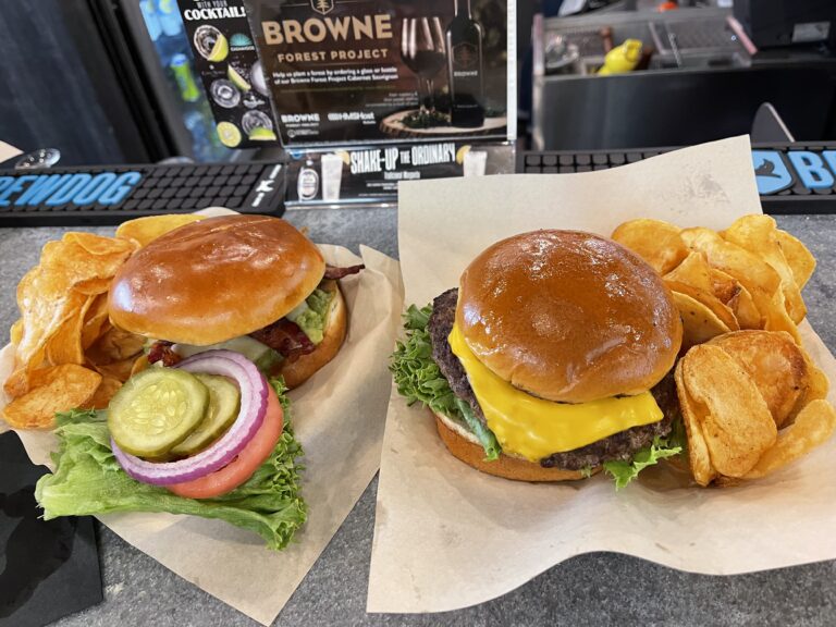 Two burgers sit next to each other on the top of a bar. On the left, a cheeseburger with toppings. On the right, a grilled chicken sandwich with bacon and potato chips.
