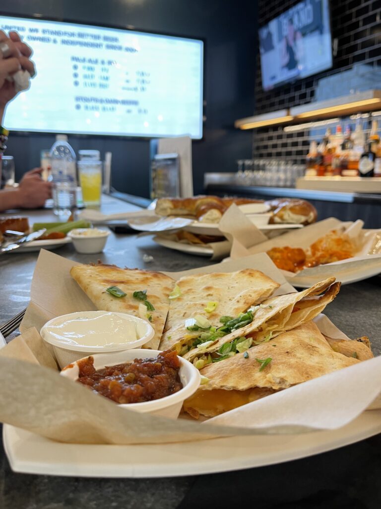 A quesadilla sits on a plate with two types of dipping sauces. In the background is a well-lit board with menu items, and other plates that have been picked at.