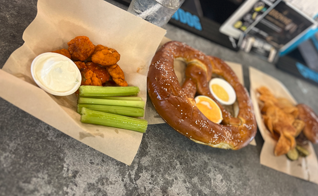 Buffalo chicken bites sit in a basket with blue cheese dip and celery. A large pretzel is beside it in another basket.