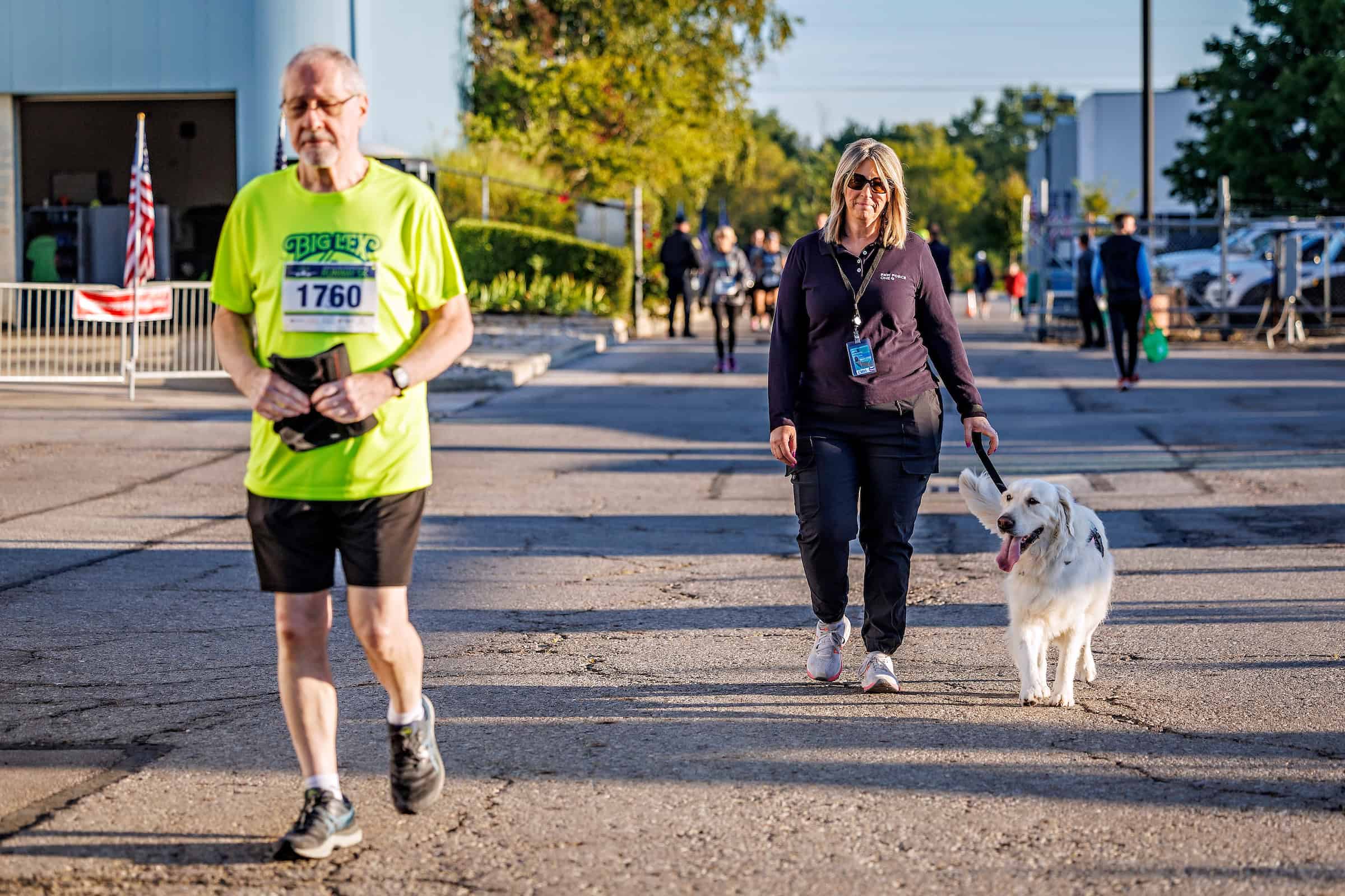 A 5K racer jogs past as a woman walks behind him with a white medium-sized dog on a leash.