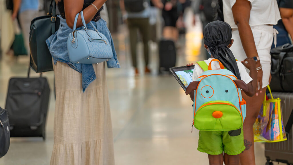 A child, along with two adults beside them, appear to be in a busy terminal, each carrying luggage, waiting for their plane to board.