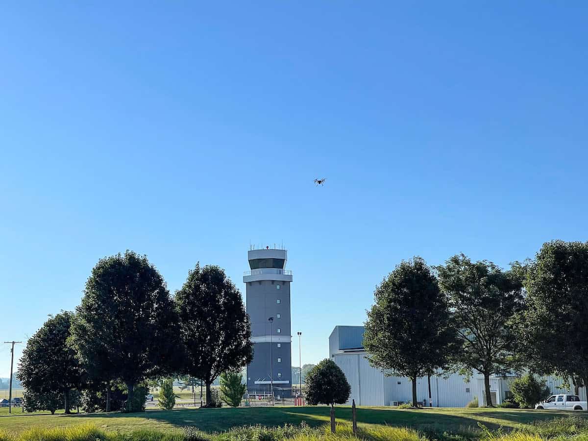 View of a sunny day at Rickenbacker International Airport featuring the control tower, surrounding buildings, lush greenery, and a drone flying overhead.
