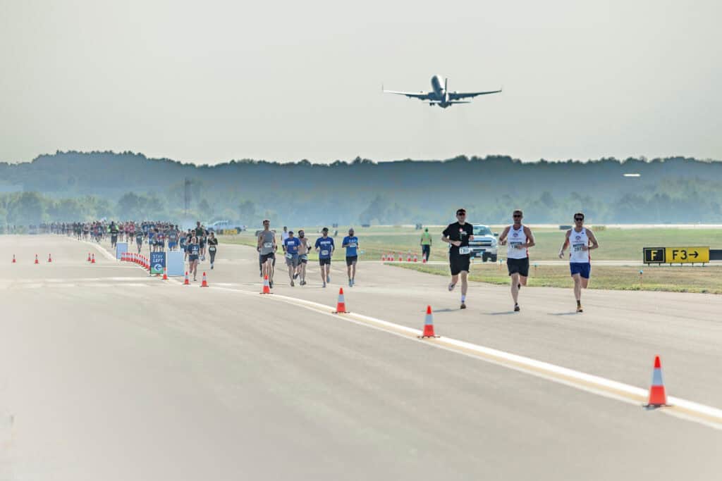 Participants running on a runway during a race event, with an airplane taking off in the background.