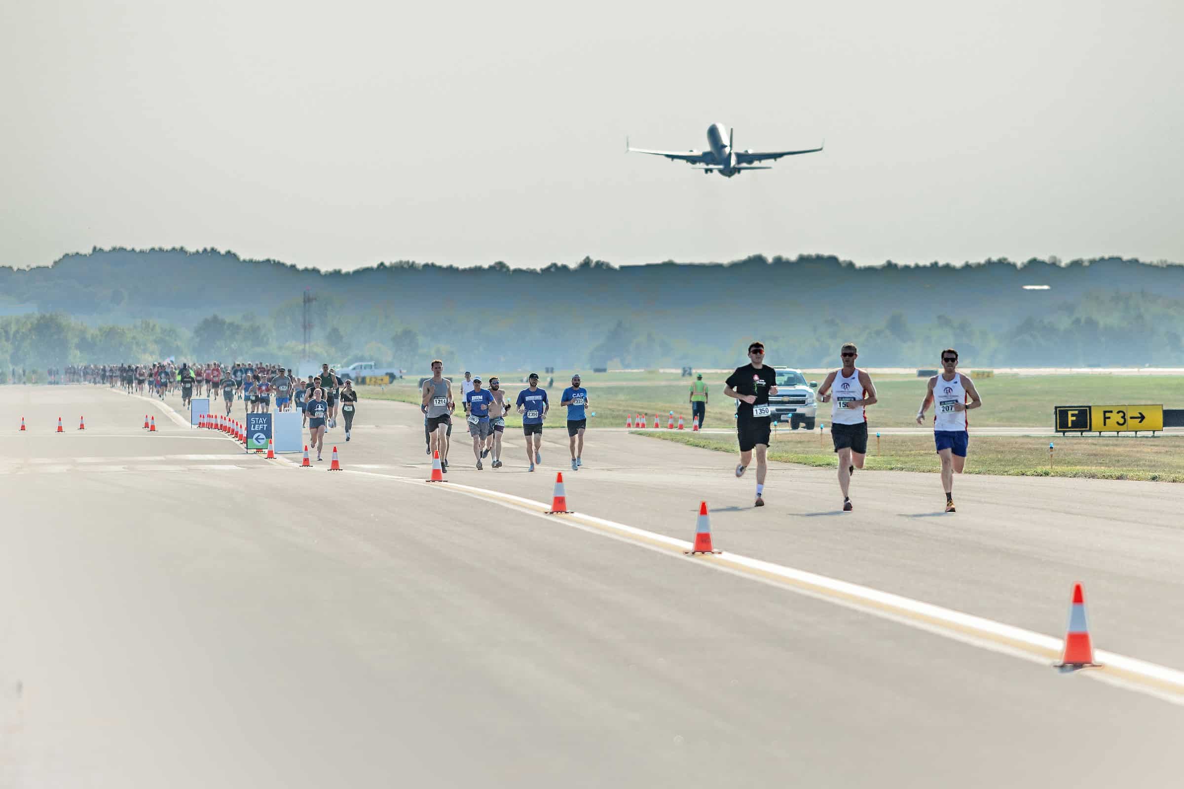 Participants running on a runway during a race event, with an airplane taking off in the background.