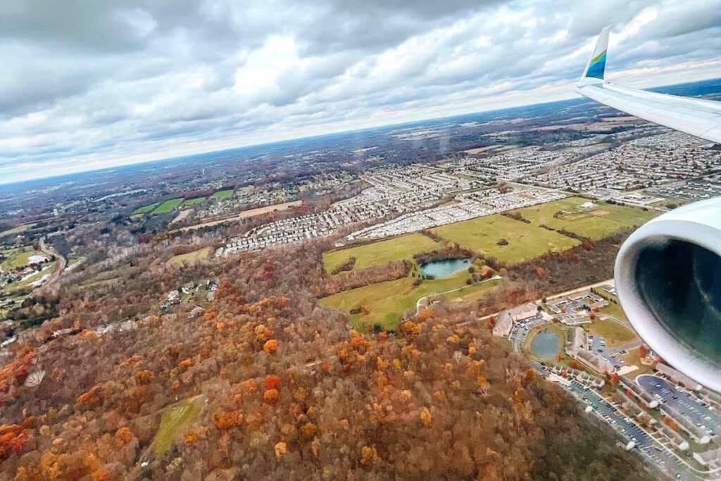 An Alaska Airlines wing flies over the suburbs of Columbus, Ohio during a fall day. Bright orange and rust colored tree tops pass below.